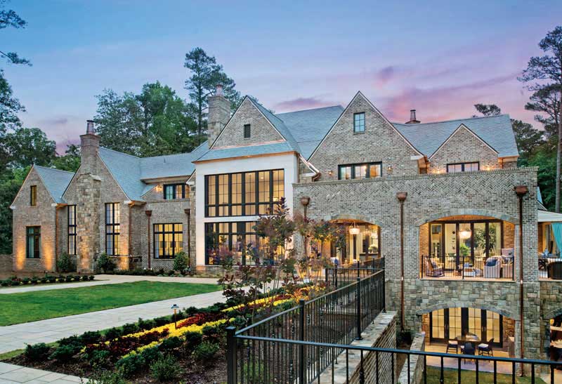 Luxurious, multi-story stone mansion with large windows, multiple gables, and a manicured garden. The house is surrounded by a patio and a fenced walkway, set against a backdrop of trees and a twilight sky.