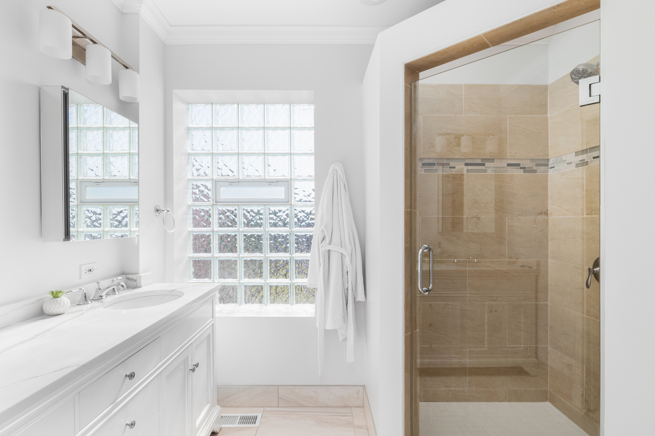 A white bathroom with a brown tiled shower. A bathroom with a white cabinet and countertop, glass block window, and a shower with brown tiles. Glass Block Window in Bathroom - Consolidated Builders Supply